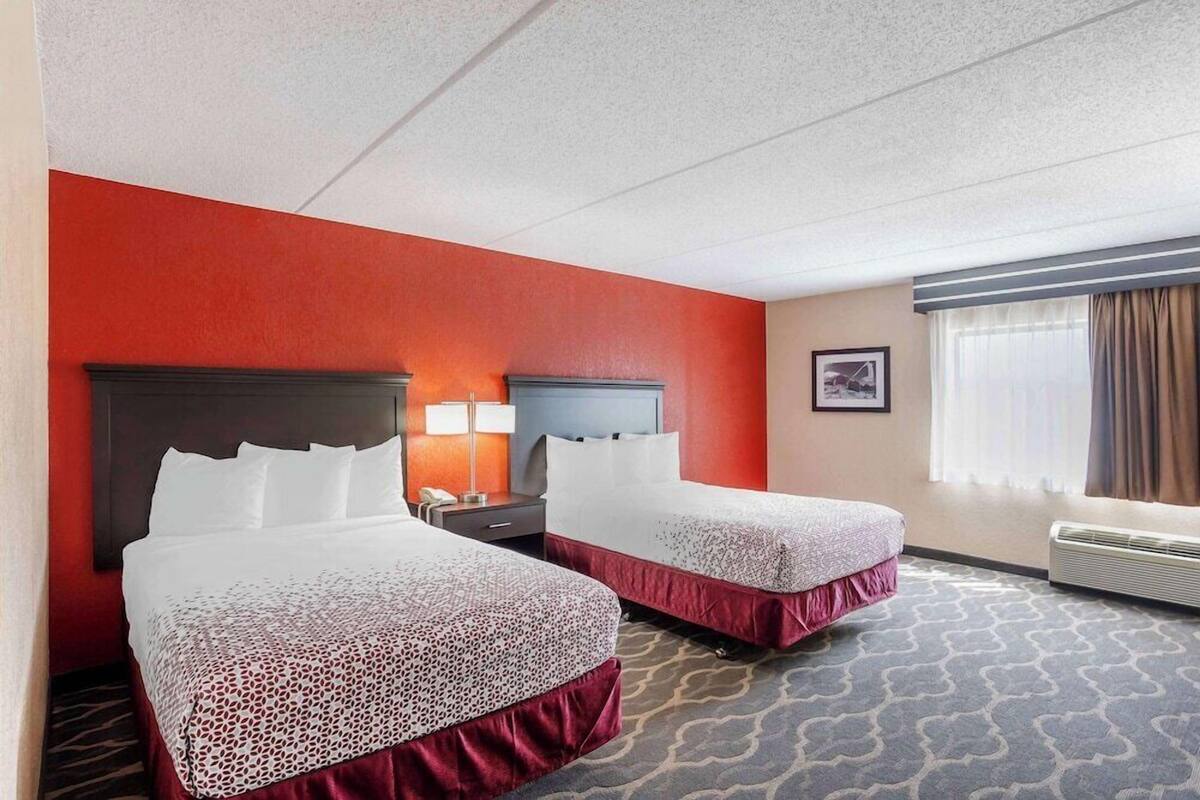 A hotel room with two beds, a red accent wall, a large window with curtains, and a carpeted floor.