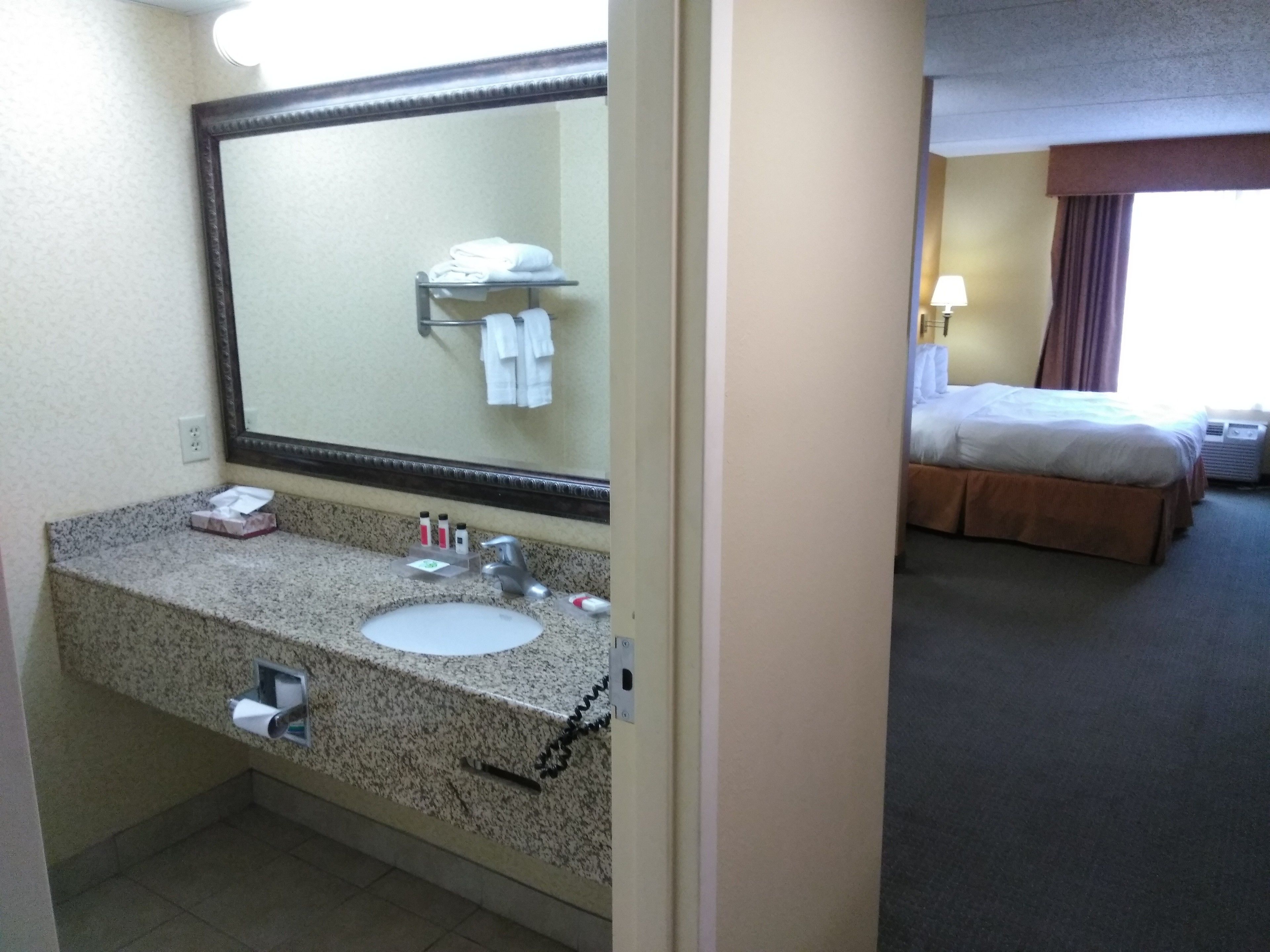 A hotel bathroom with a granite countertop sink, a large mirror, and a towel rack with folded towels.