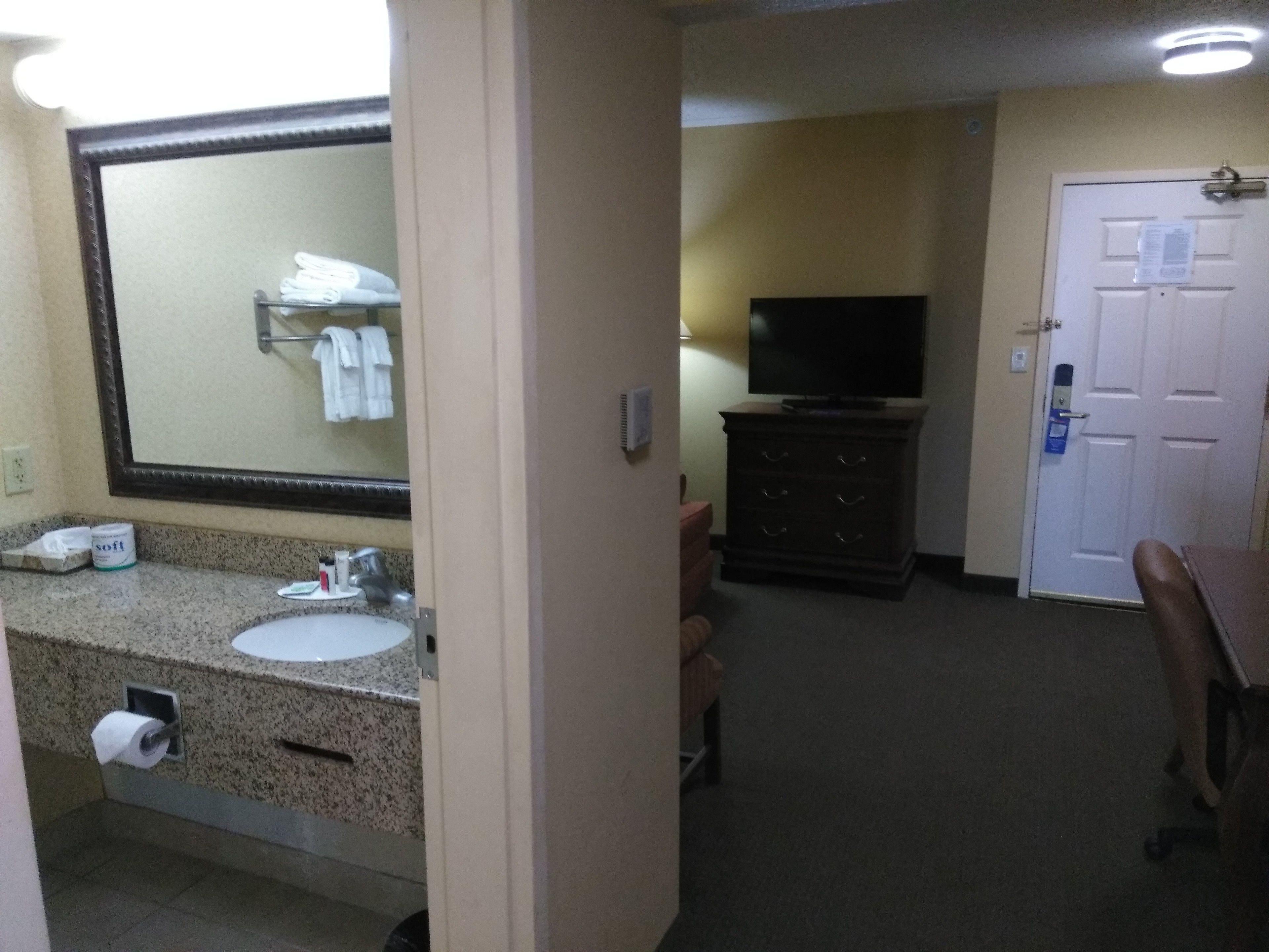 A hotel bathroom with a sink, mirror, and toiletries. There is a television and a door in the background.