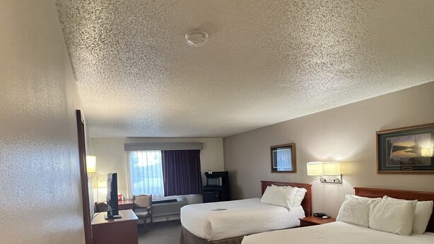 A hotel room with two beds, a desk, a chair, a TV, and a window with curtains. - Serena Inn & Suites - Sundance (Sundance)