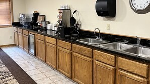 A kitchen with wooden cabinets, a black countertop, a coffee machine, a waffle maker, and a clock on the wall. - Serena Inn & Suites - Sundance (Sundance)