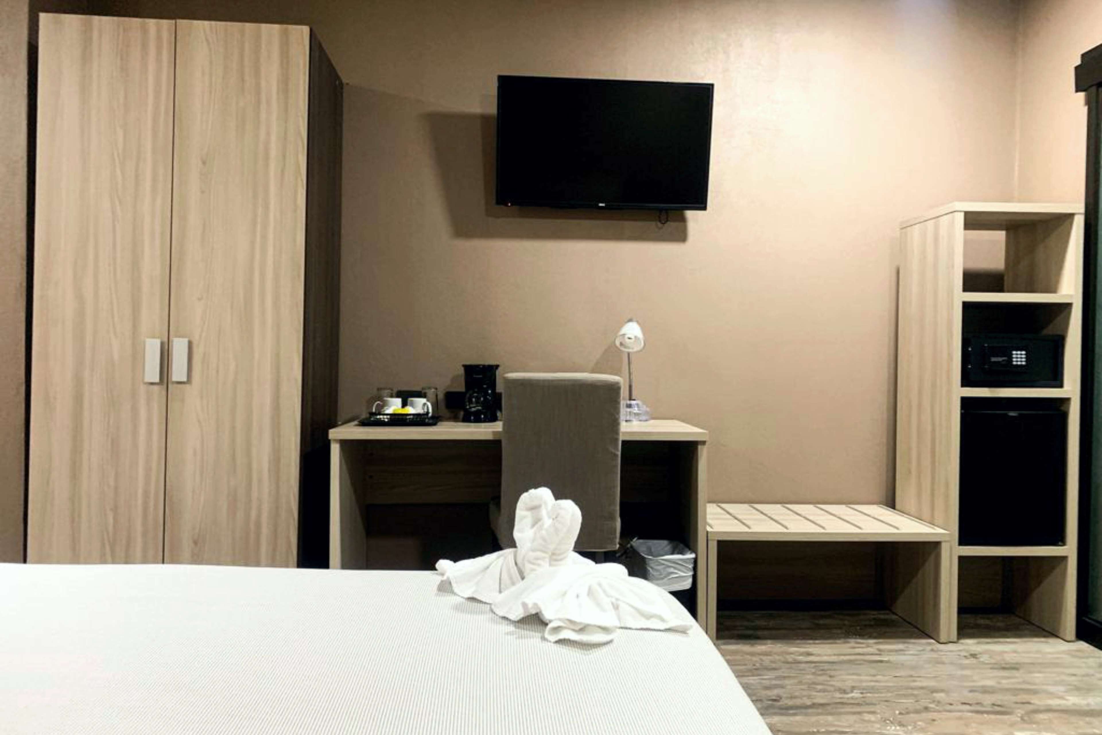 A hotel room with a bed, a desk with a chair, a TV mounted on the wall, and a wardrobe.