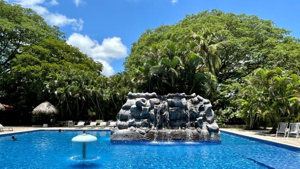 A swimming pool with a rock waterfall feature surrounded by lush greenery. - Best Western El Sitio Hotel (Liberia)