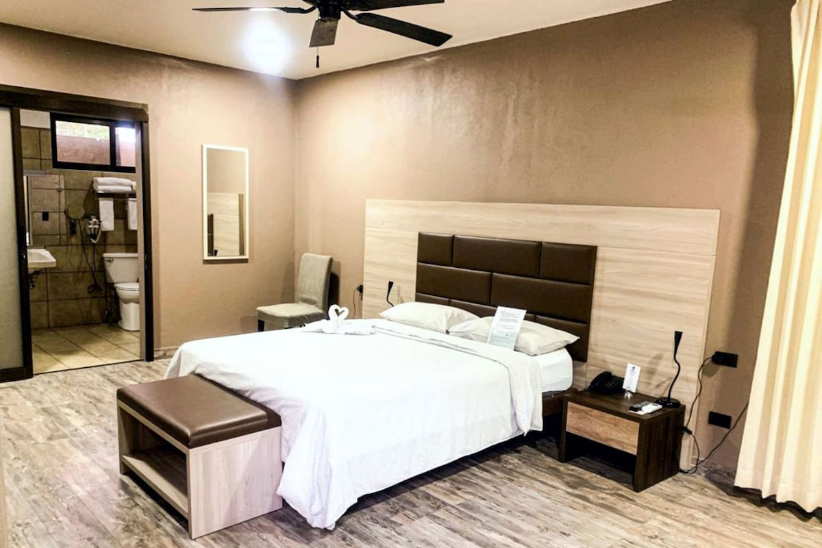 Habitación de hotel con cama, mesitas de noche, espejo, ventilador de techo y baño visible a través de una puerta abierta.