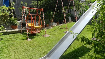 Children can play on the slide and swings in this outdoor playground area surrounded by lush greenery.