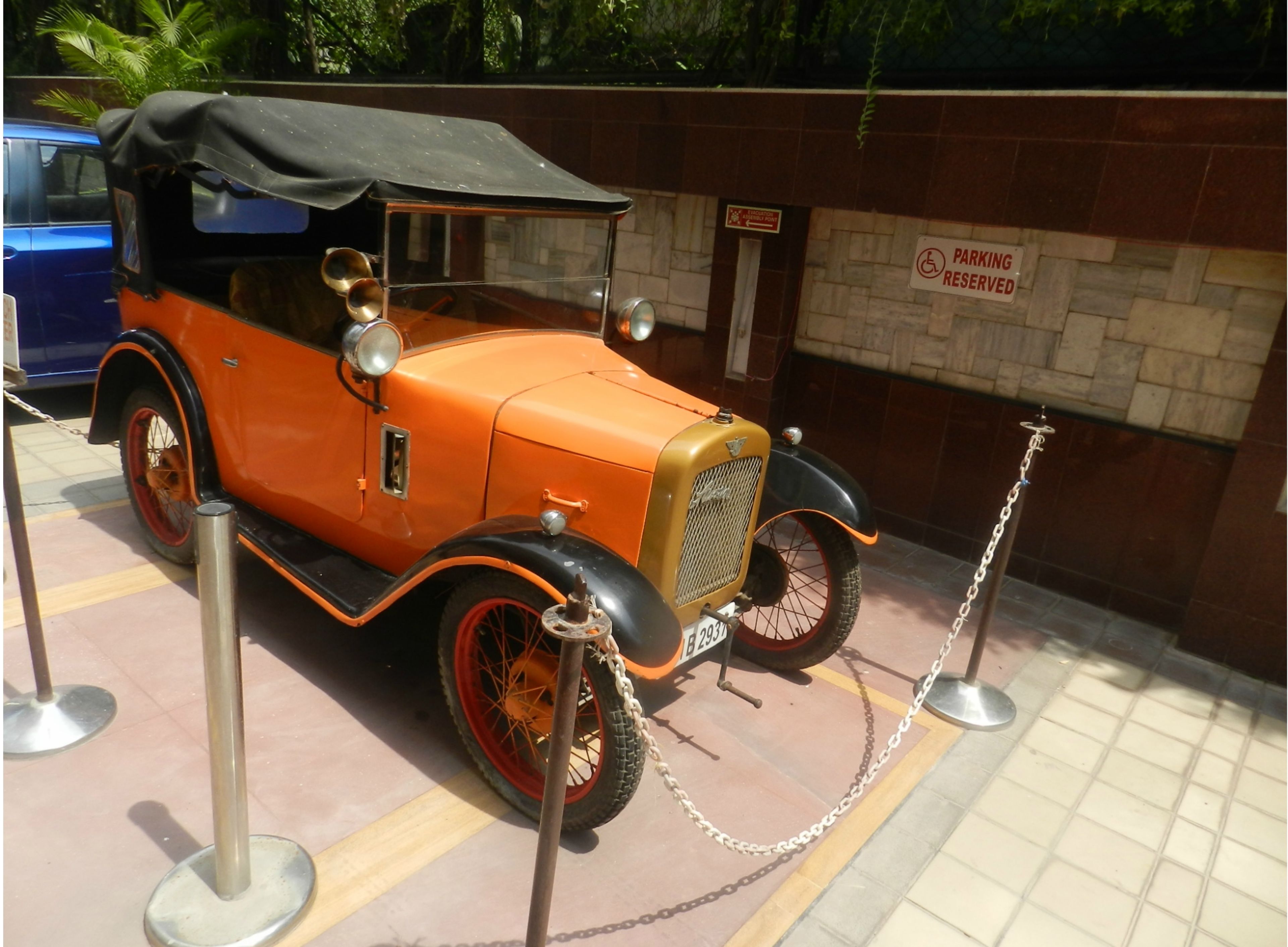 Un coche antiguo exhibido, rodeado por una barrera de cadena, con un cartel de 'PARKING RESERVED' al fondo.