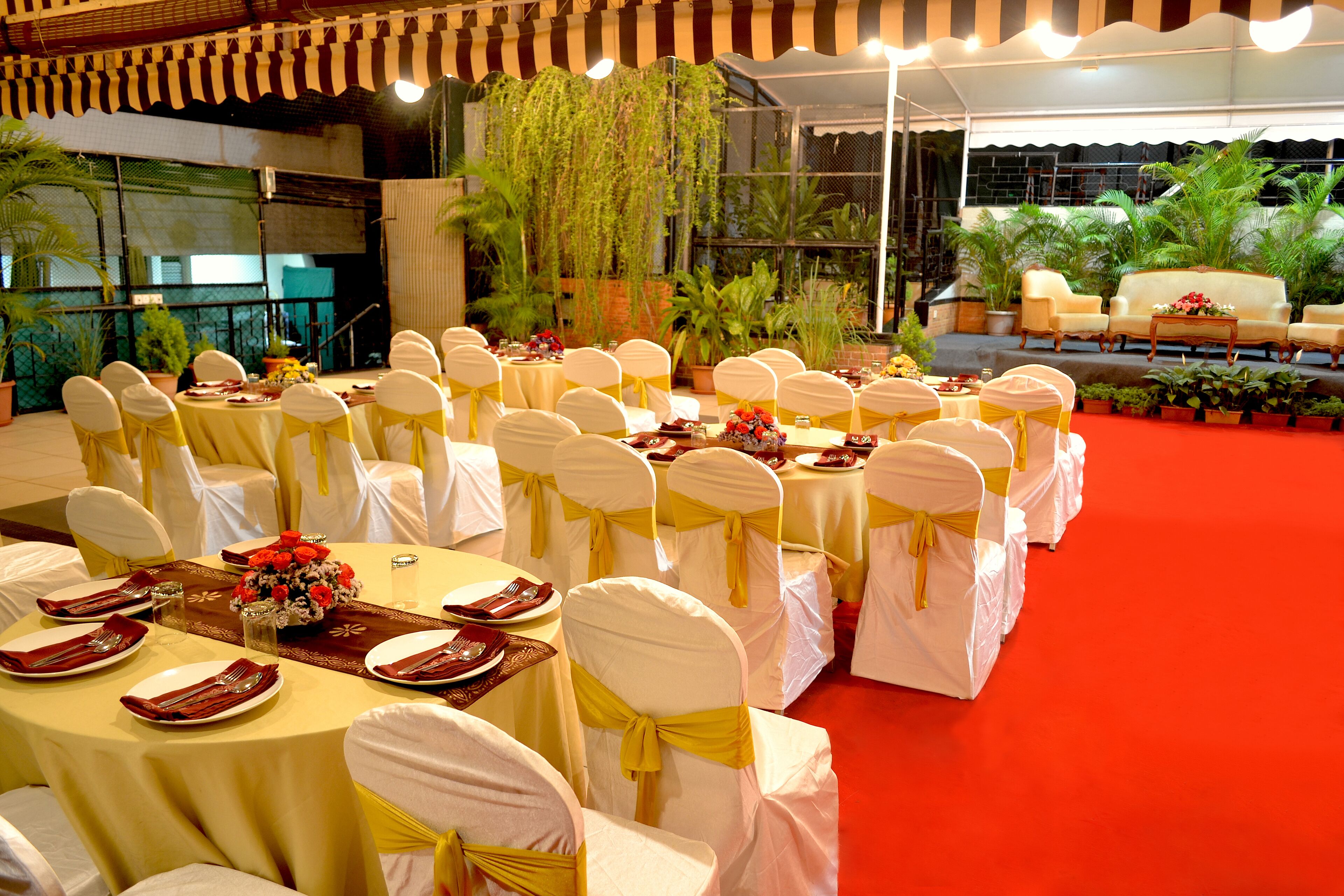 A banquet hall with round tables, white chairs with yellow sashes, and a stage with sofas and potted plants.