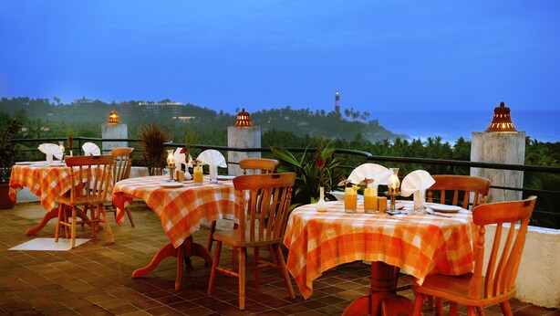 An outdoor dining area with checkered tablecloths and wooden chairs, overlooking a coastal landscape with hills and a lighthouse. - Swagath Holiday Resort (Neyyattinkara)