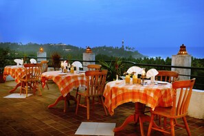 Un área de comedor al aire libre con manteles a cuadros y sillas de madera, con vistas a un paisaje costero con colinas y un faro.
