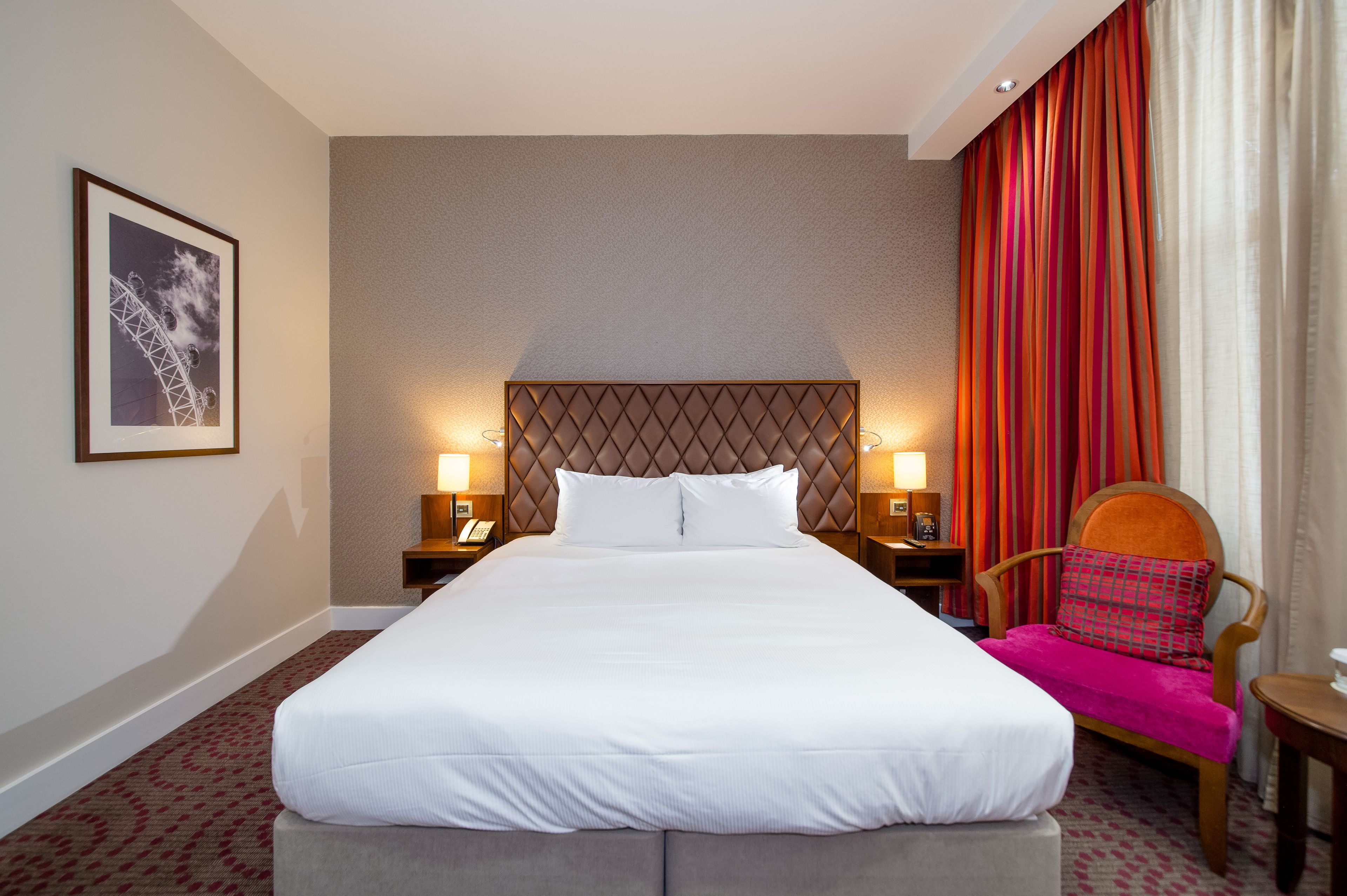 A hotel room with a large bed, two bedside tables with lamps, a chair with a colorful cushion, and a framed picture on the wall.