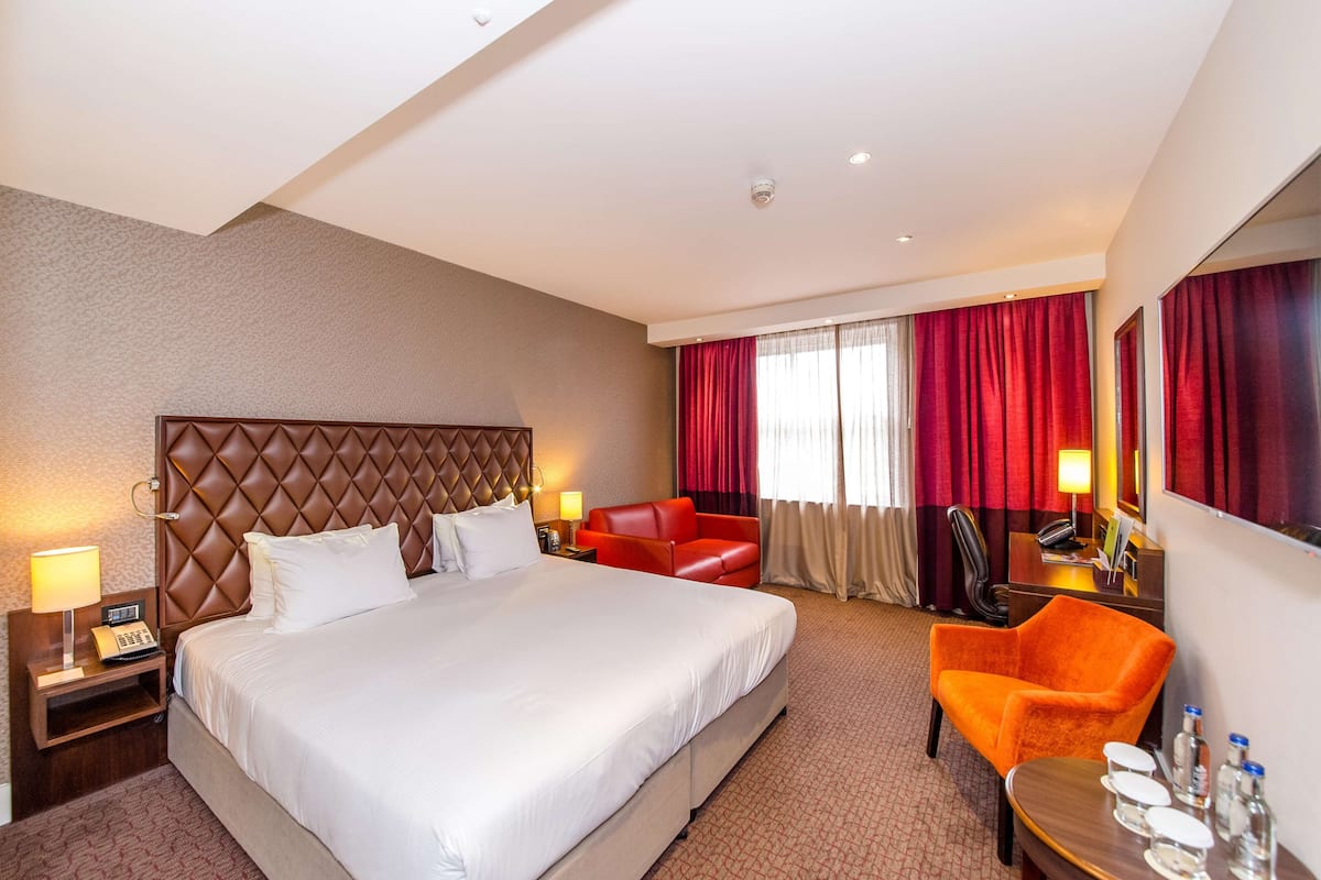 A hotel room with a large bed, a red armchair, a wooden nightstand with a lamp, and a small table with bottles.