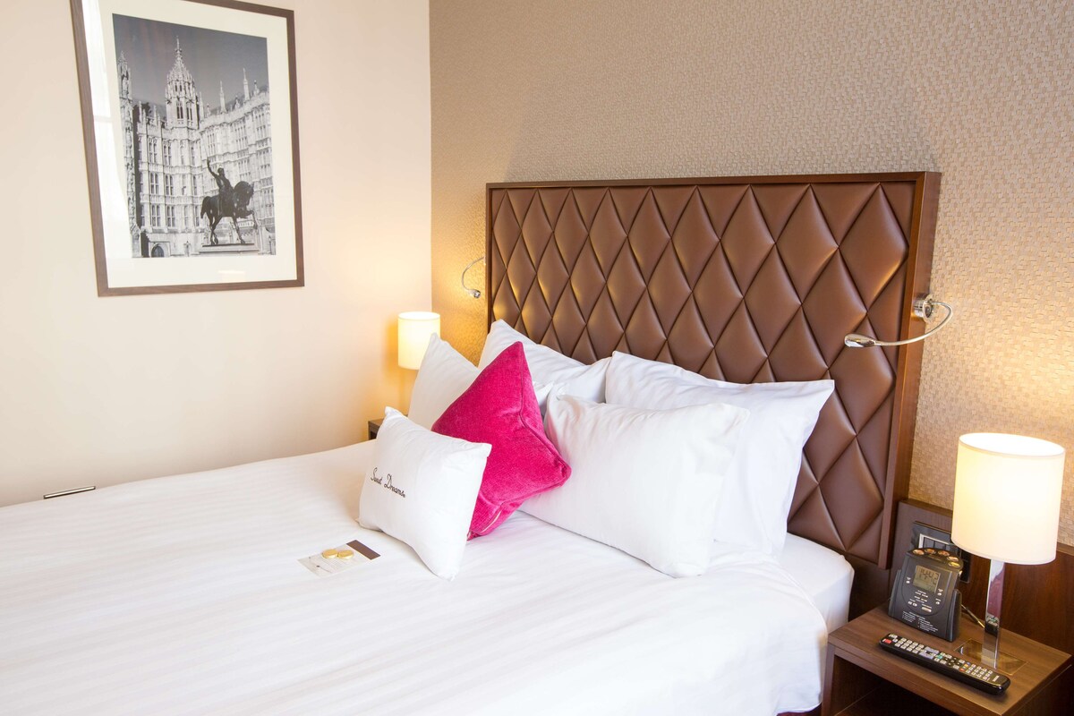 A hotel room with a large bed, a headboard, two bedside lamps, a framed picture on the wall, and a remote control on the nightstand.