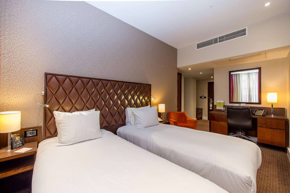 A hotel room with two beds, a desk, a chair, and a window with curtains.