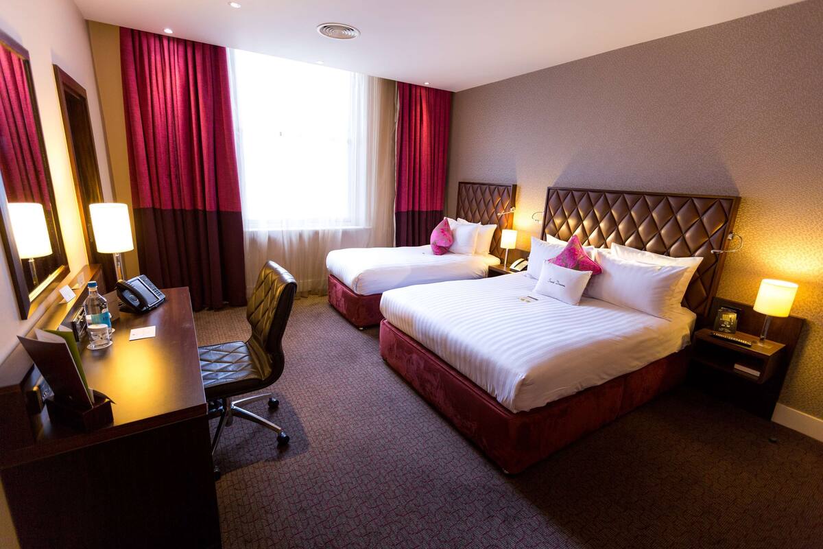A hotel room with two beds, a desk with a chair, a desk lamp, a telephone, and a window with curtains.