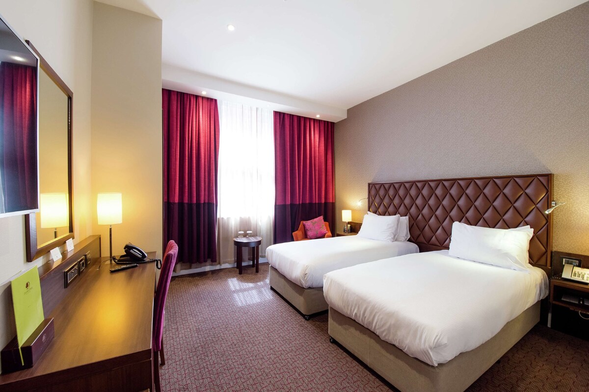 A hotel room with two beds, a desk with a telephone, a mirror, and a small table with a lamp.