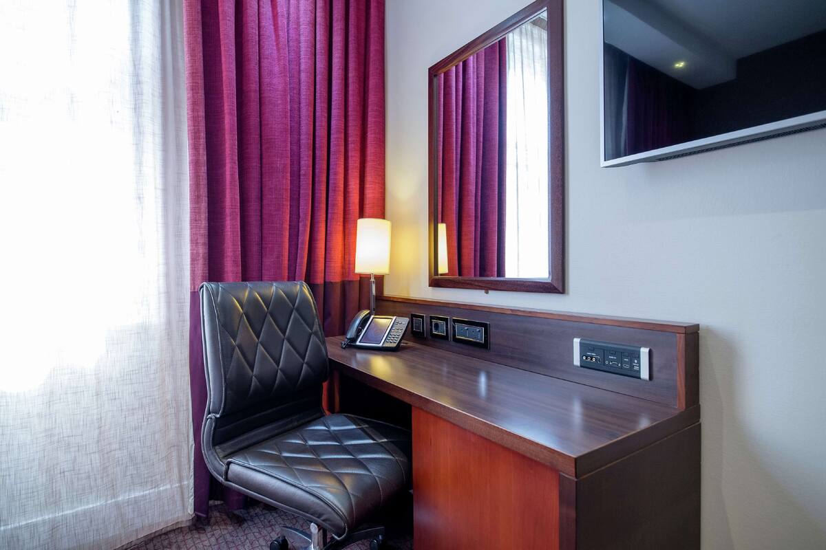 A hotel room with a leather chair, a desk with a telephone, a mirror, and a window with curtains.