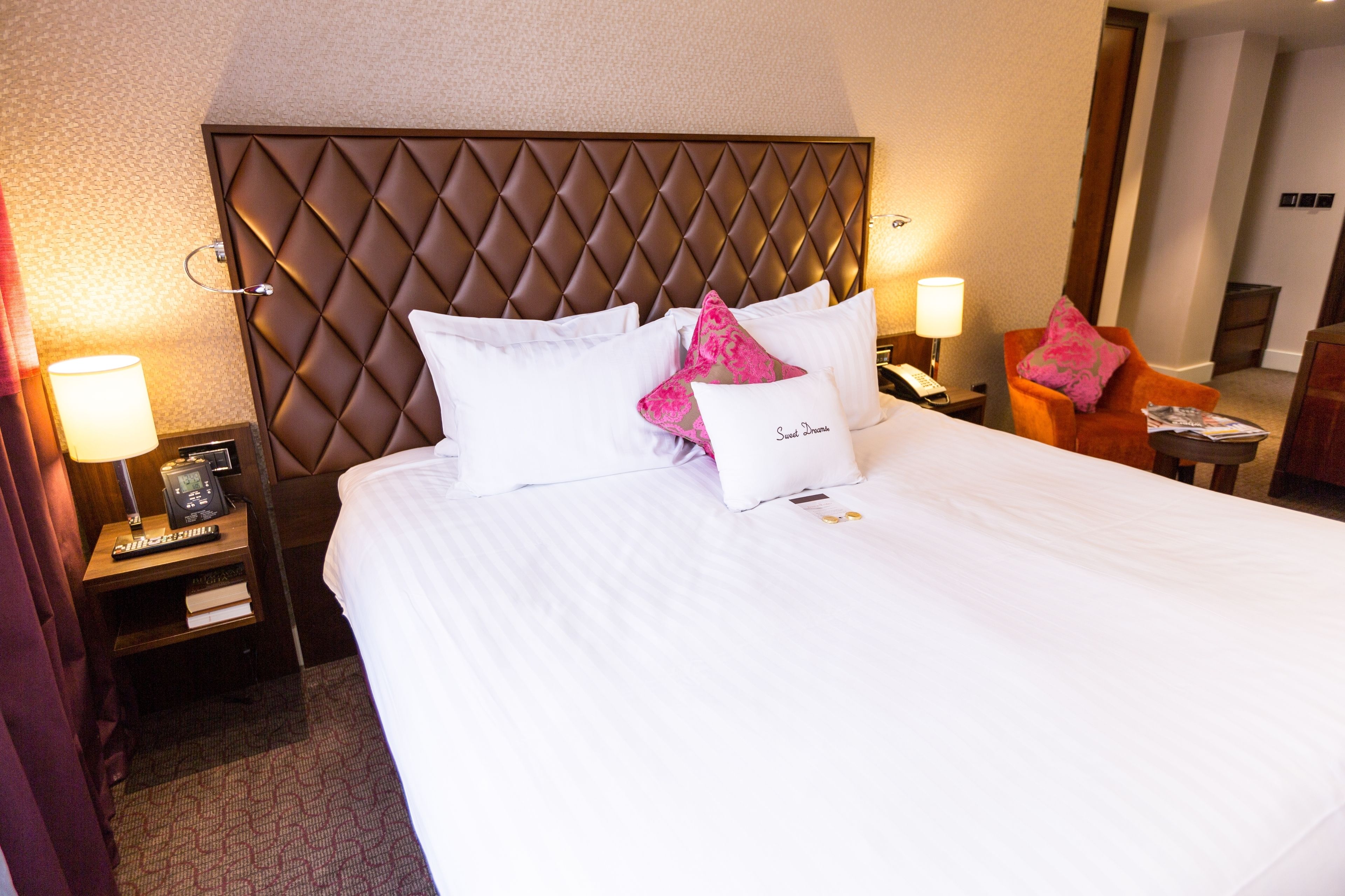 A hotel room with a large bed, a headboard with a diamond pattern, a nightstand with a lamp, and a chair with a cushion.