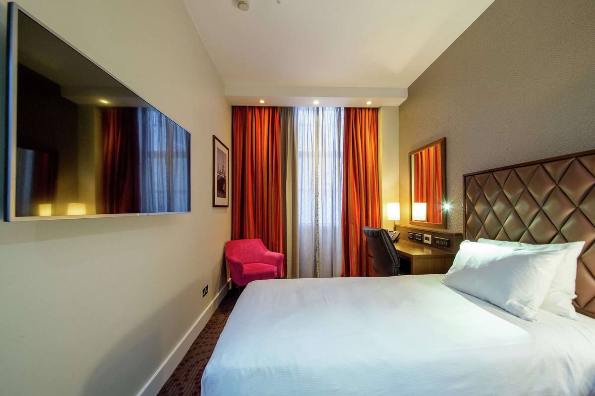 A hotel room with a large bed, a desk with a chair, a TV mounted on the wall, and a window with curtains.
