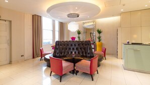 A modern hotel room with a round table, red upholstered chairs, a tufted sofa, a chandelier, a large mirror, and a green kitchen counter. - Doubletree by Hilton London Marble Arch (London)