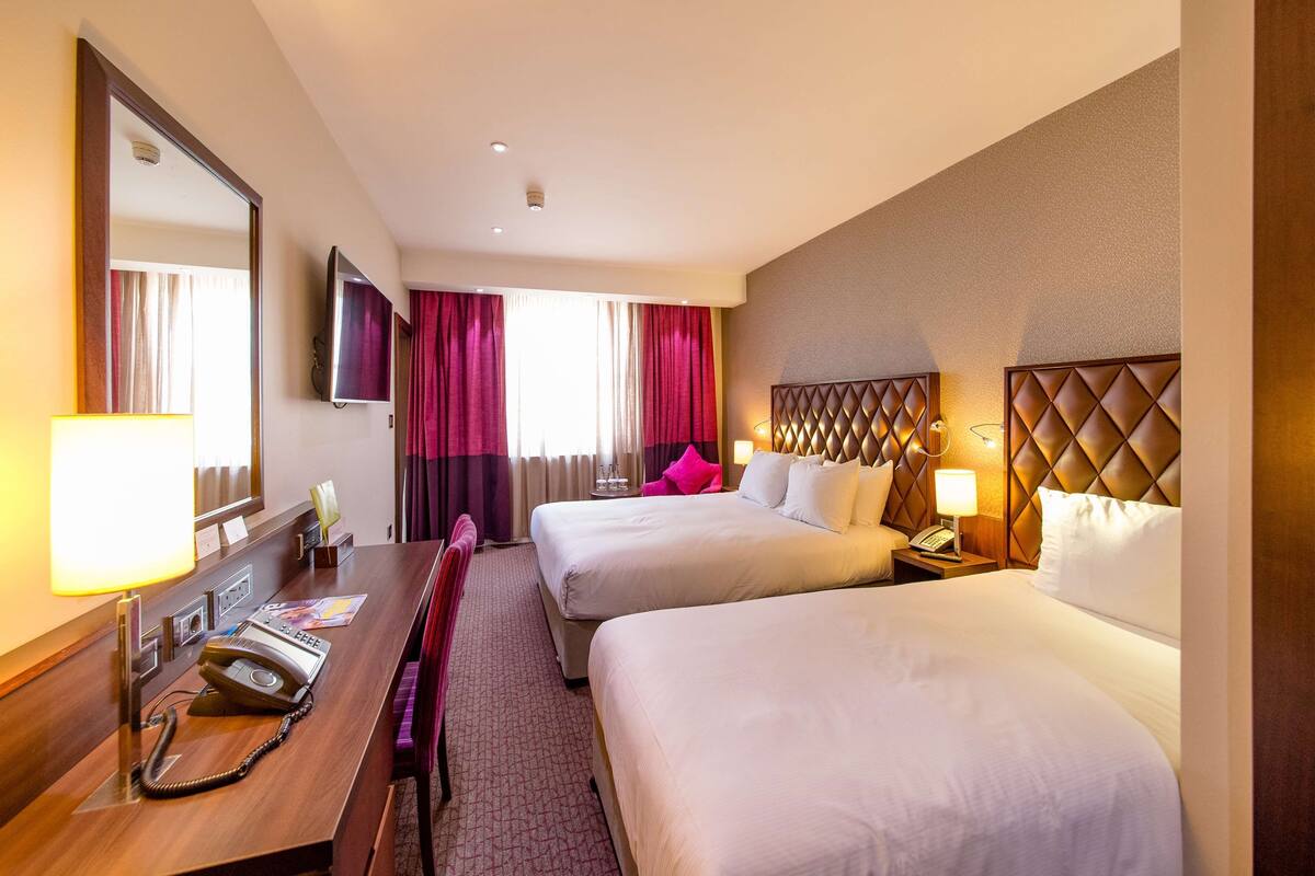 A hotel room with two beds, a desk with a telephone, a mirror, and a television on the wall.