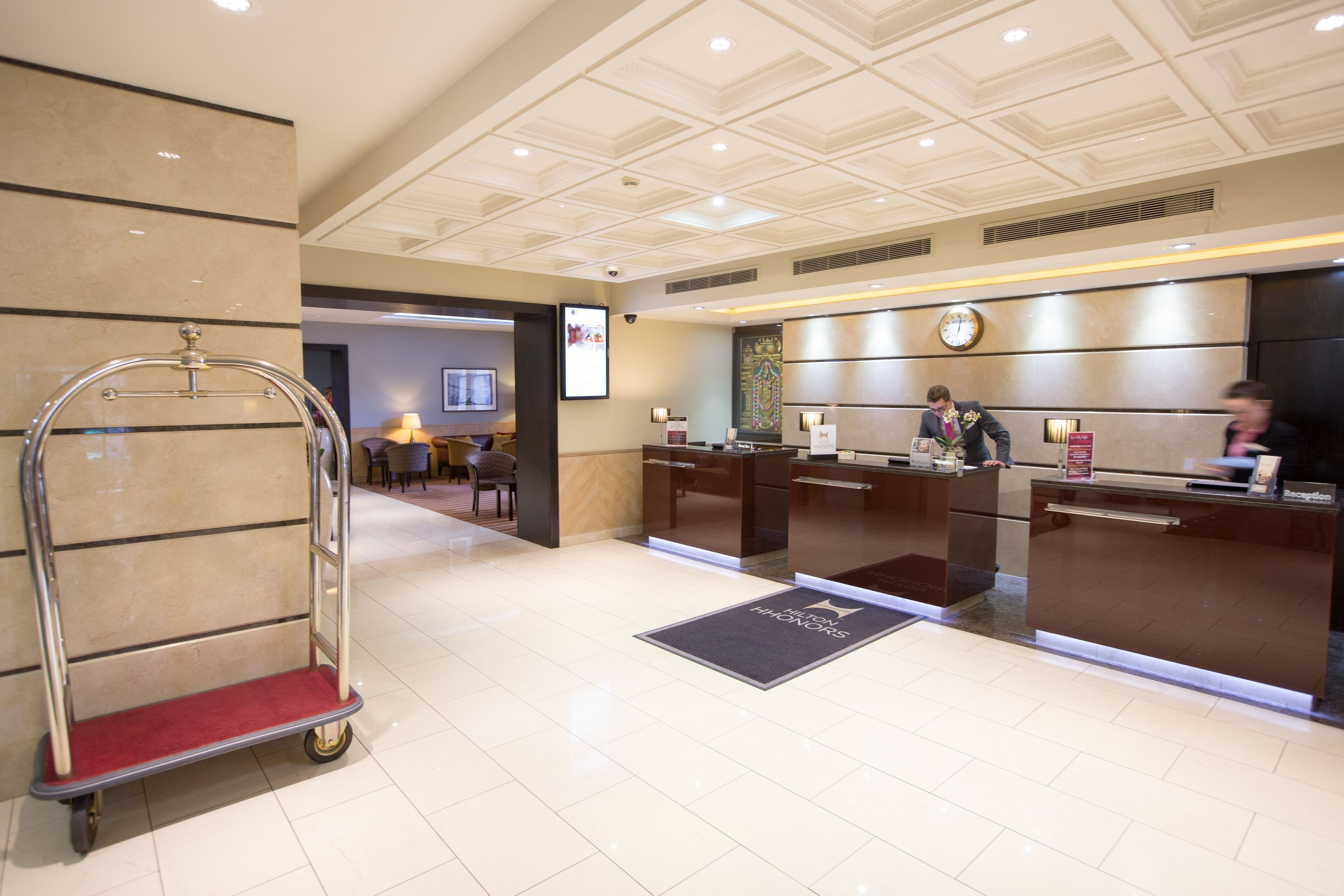 A hotel reception area with a reception desk, a clock, and a luggage cart.