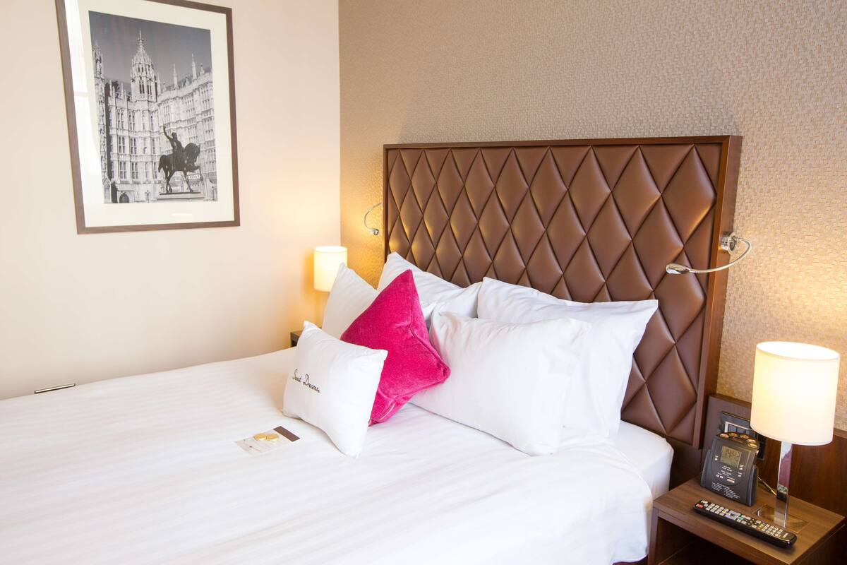 A hotel room with a large bed, a headboard, two bedside lamps, a framed picture on the wall, and a remote control on the nightstand.