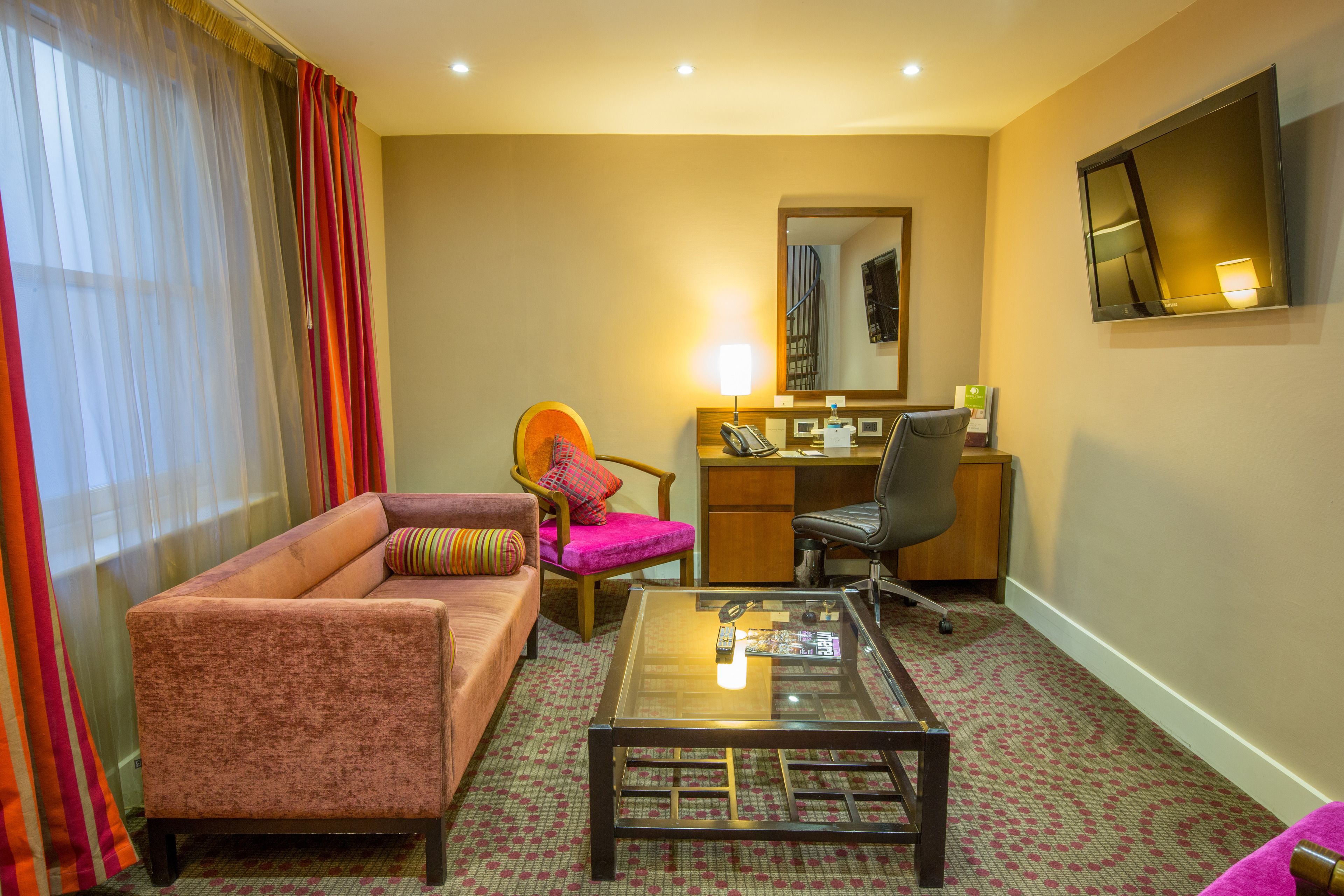 A hotel room with a sofa, armchair, desk, chair, and a TV mounted on the wall.