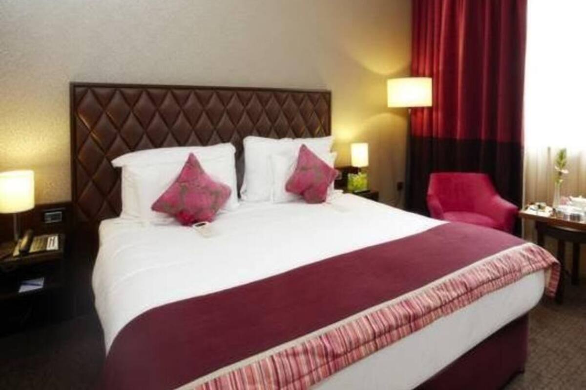 A hotel room with a large bed, a headboard, two bedside tables with lamps, a red curtain, and a chair.