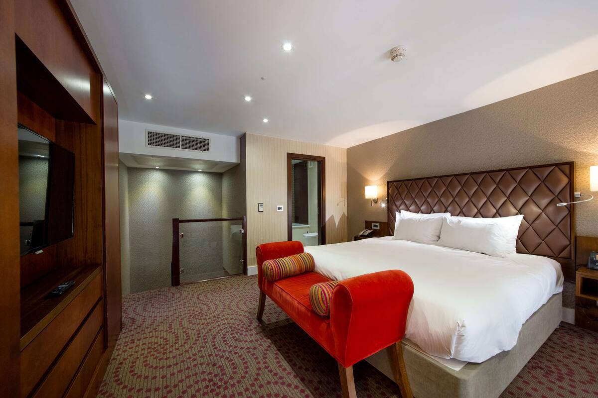 A hotel room with a large bed, a red bench, a television, a minibar, and a bathroom with a mirror and toilet.