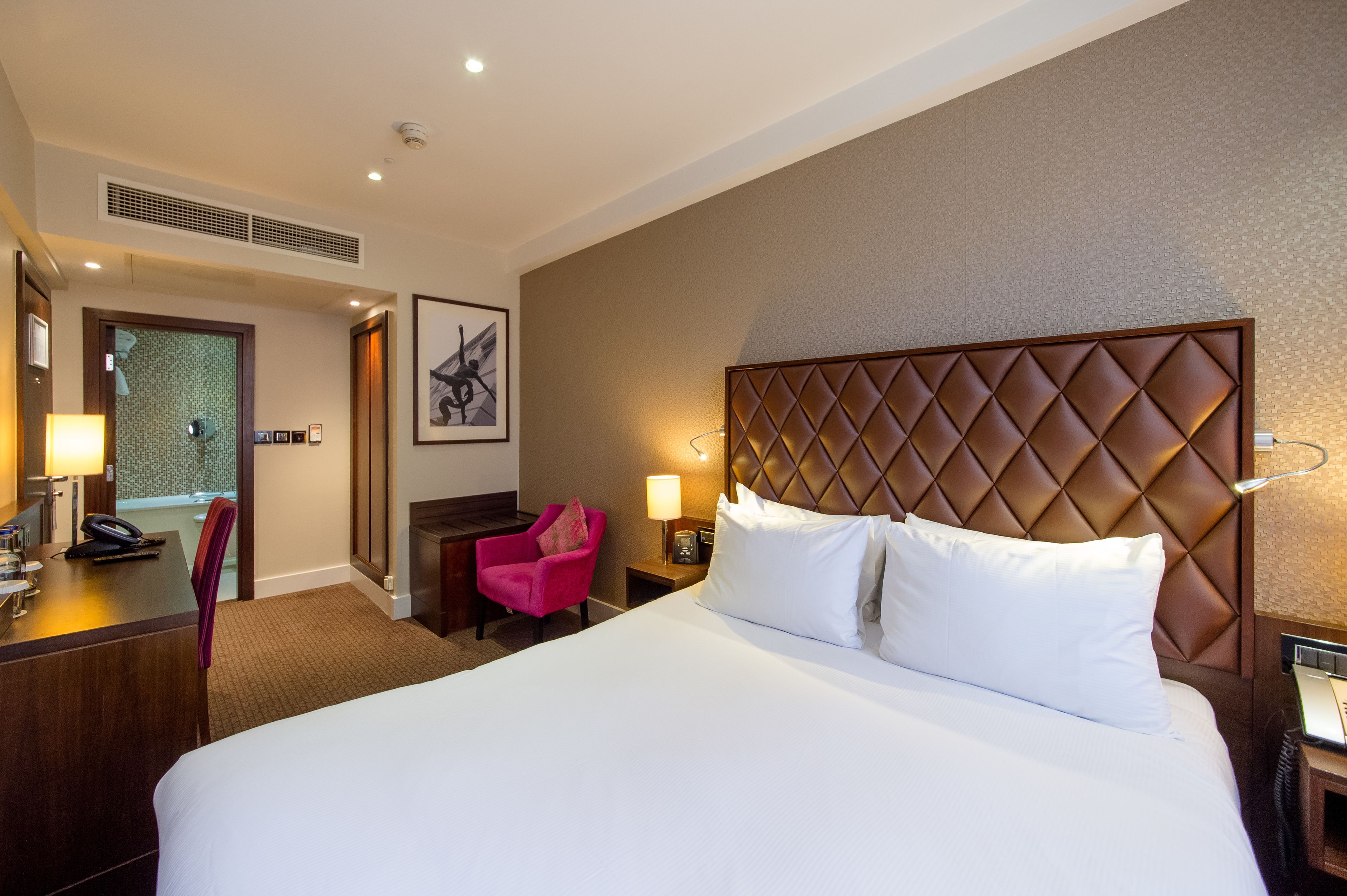 A hotel room with a large bed, a desk with a telephone, a chair, a nightstand with a lamp, and a wall-mounted artwork.