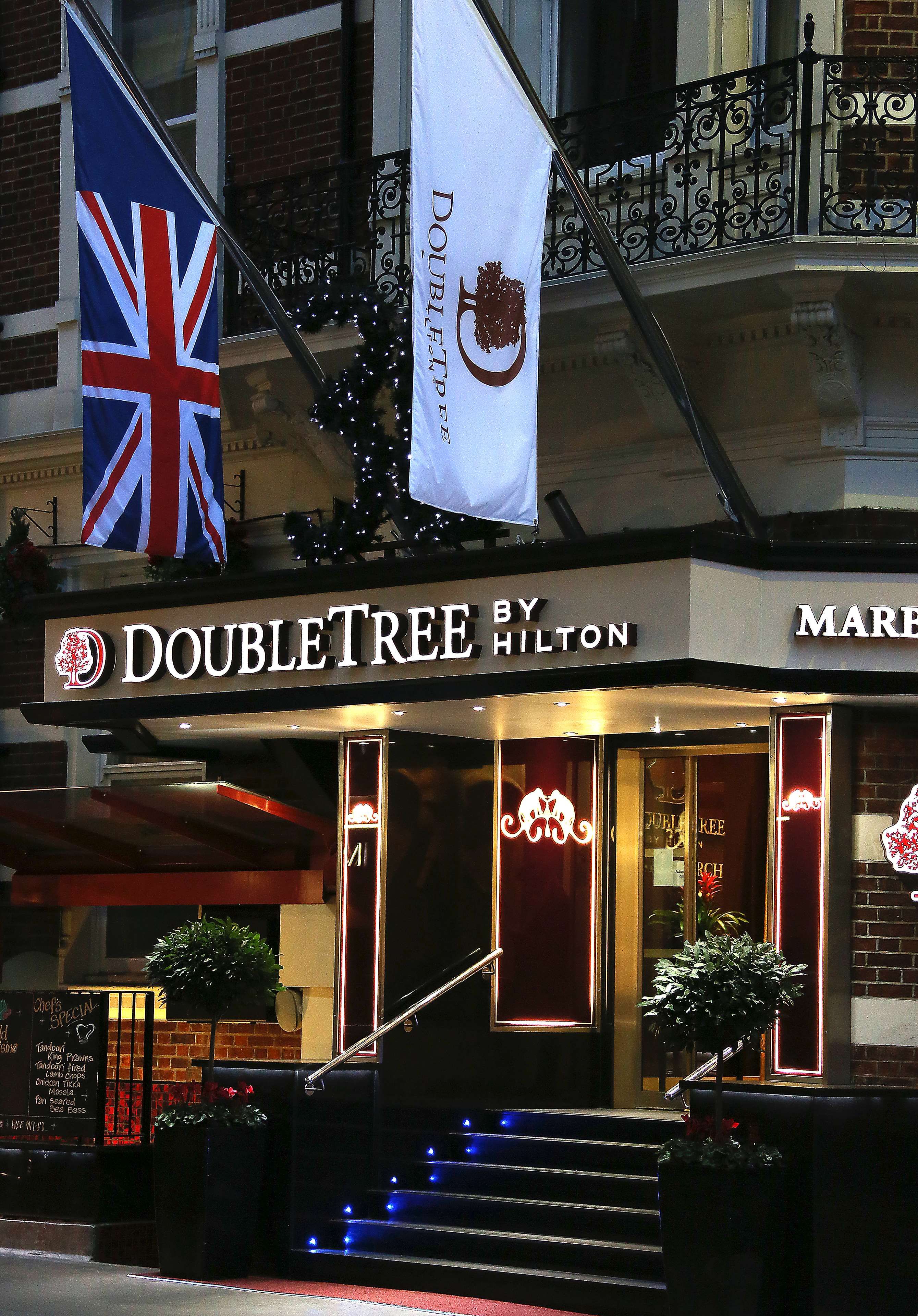 A DoubleTree by Hilton hotel entrance with a flag, illuminated sign, and steps leading up to the door.