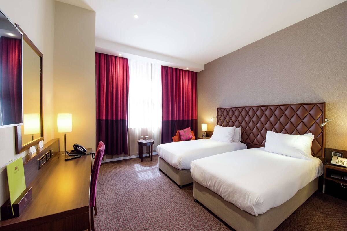A hotel room with two beds, a desk with a telephone, a mirror, and a small table with a lamp.