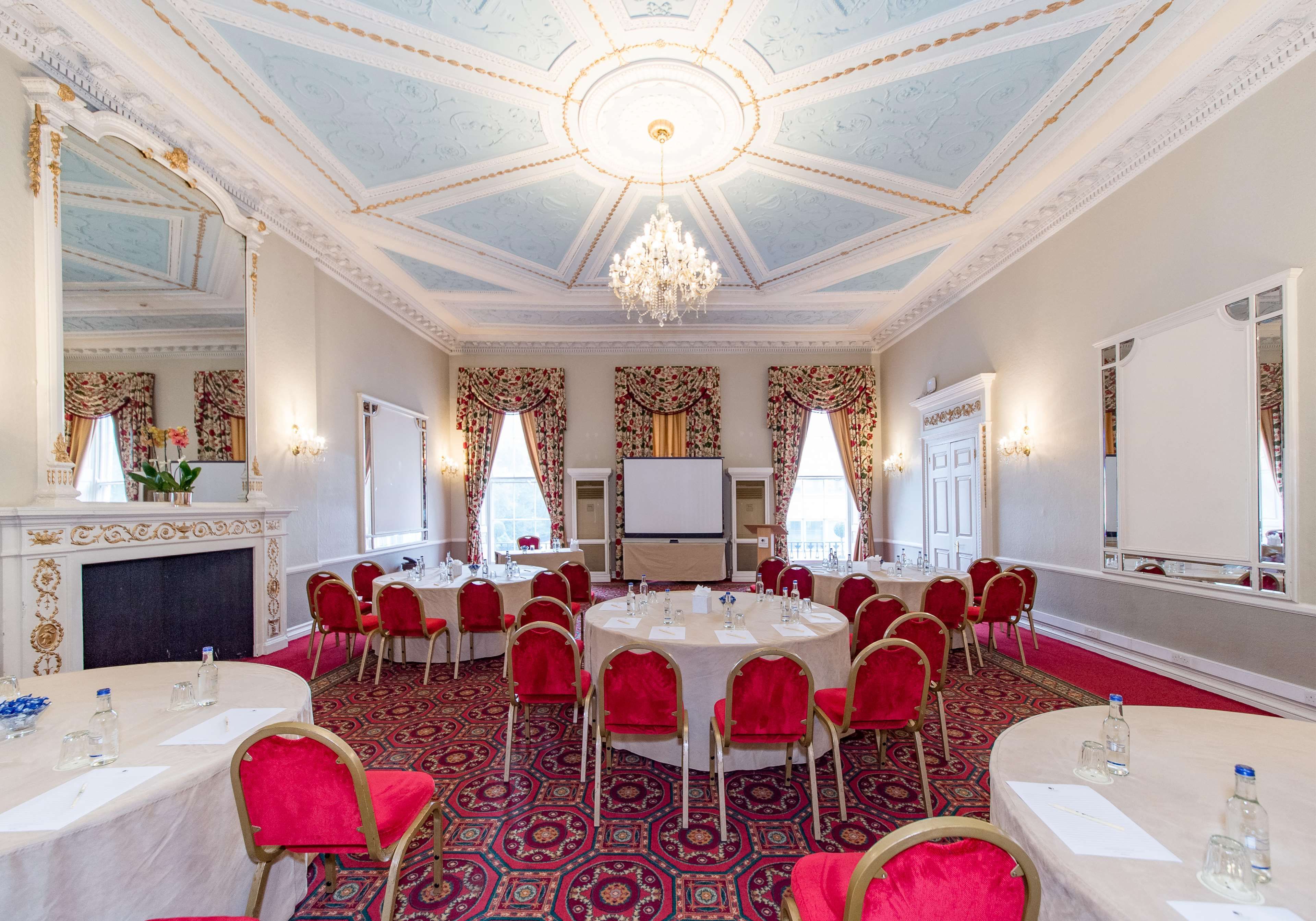 A formal conference room with round tables, red chairs, a chandelier, and ornate ceiling details.