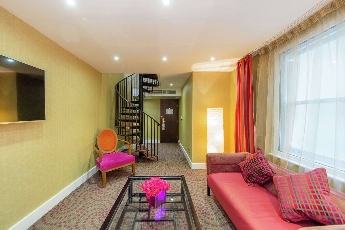 A living room with a sofa, a chair, a coffee table with flowers, a TV, a staircase, and a window with curtains.