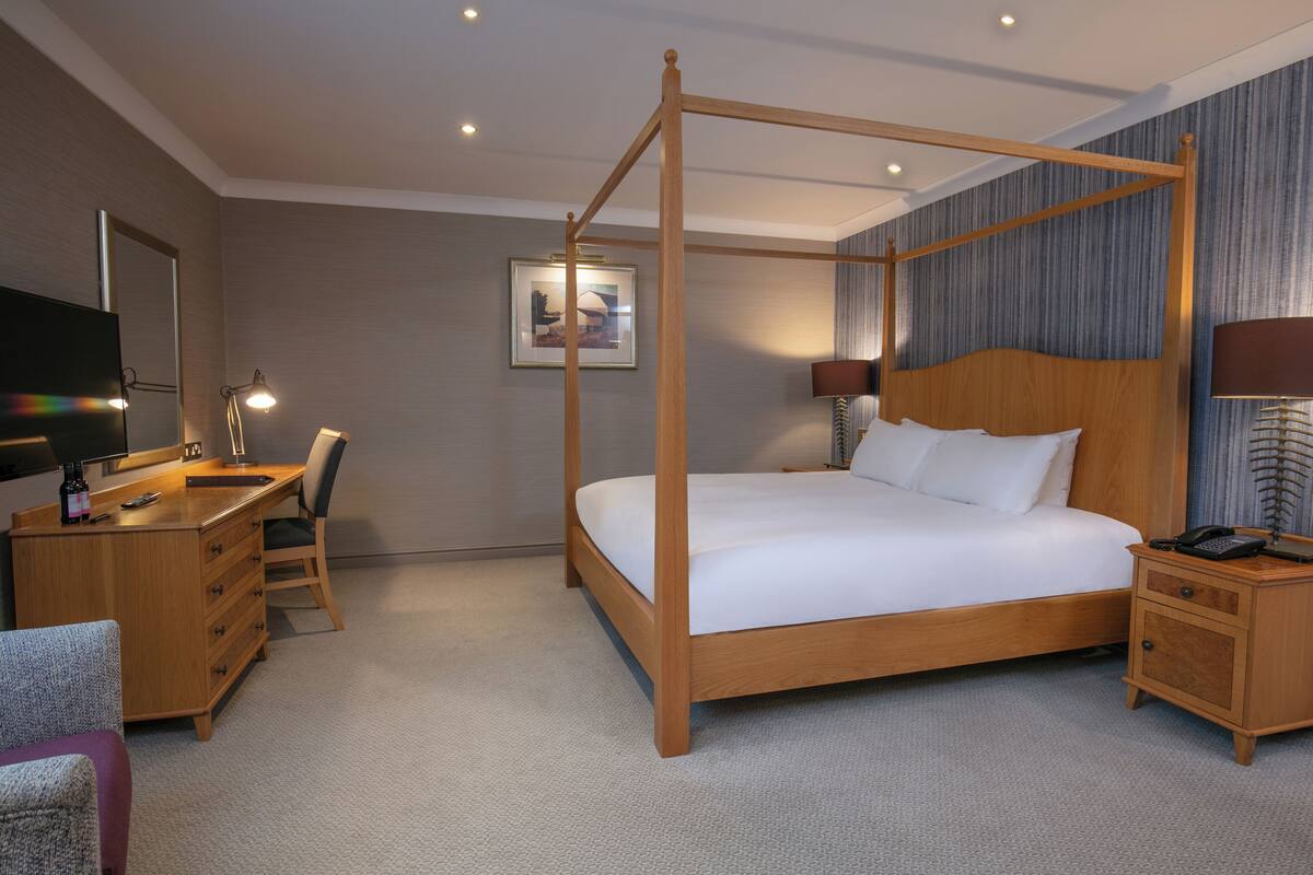 A four-poster bed with a wooden frame, a desk with a chair, a nightstand, and a television on the left side.