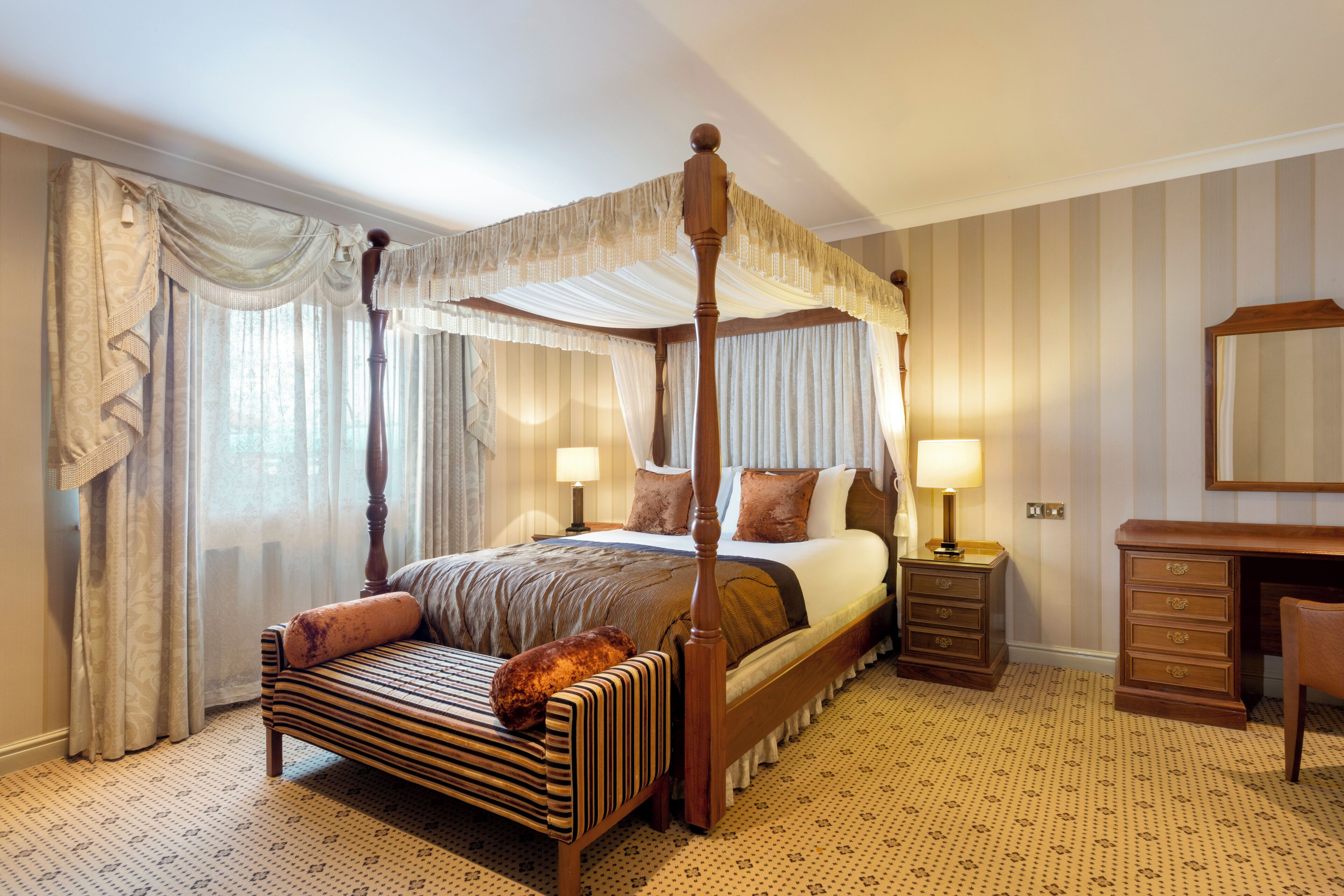 Un lit à baldaquin à quatre poteaux avec une structure en bois, un banc avec un coussin, une table de chevet avec une lampe et une commode avec des tiroirs.