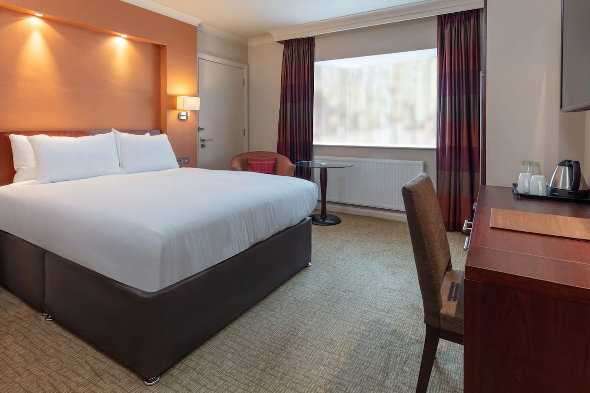 A hotel room with a large bed, a desk with a chair, a small table, and a window with curtains.