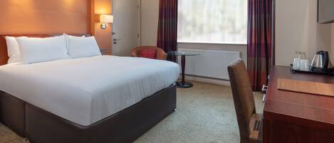A hotel room with a large bed, a desk with a chair, a small table, and a window with curtains.