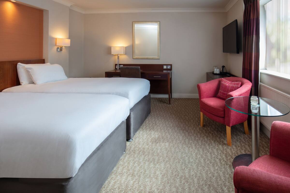 A hotel room with two beds, a desk, a chair, a TV, and a window with curtains.