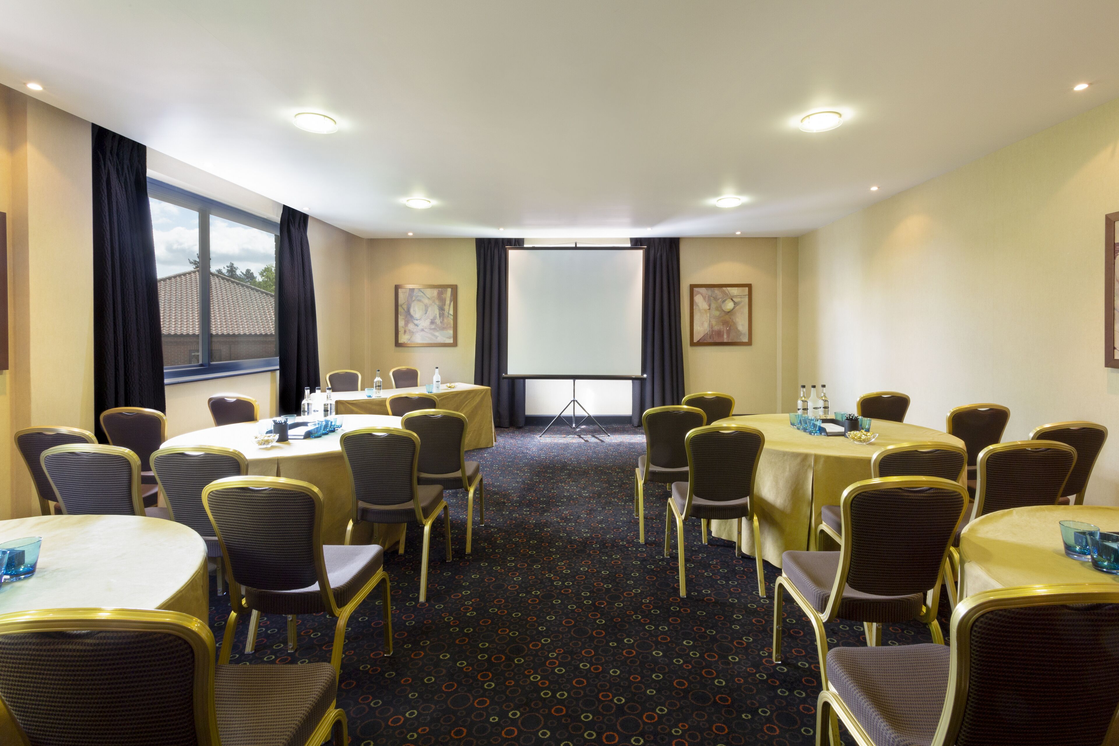 Un salón de conferencias con mesas redondas, sillas y una pantalla para proyector.