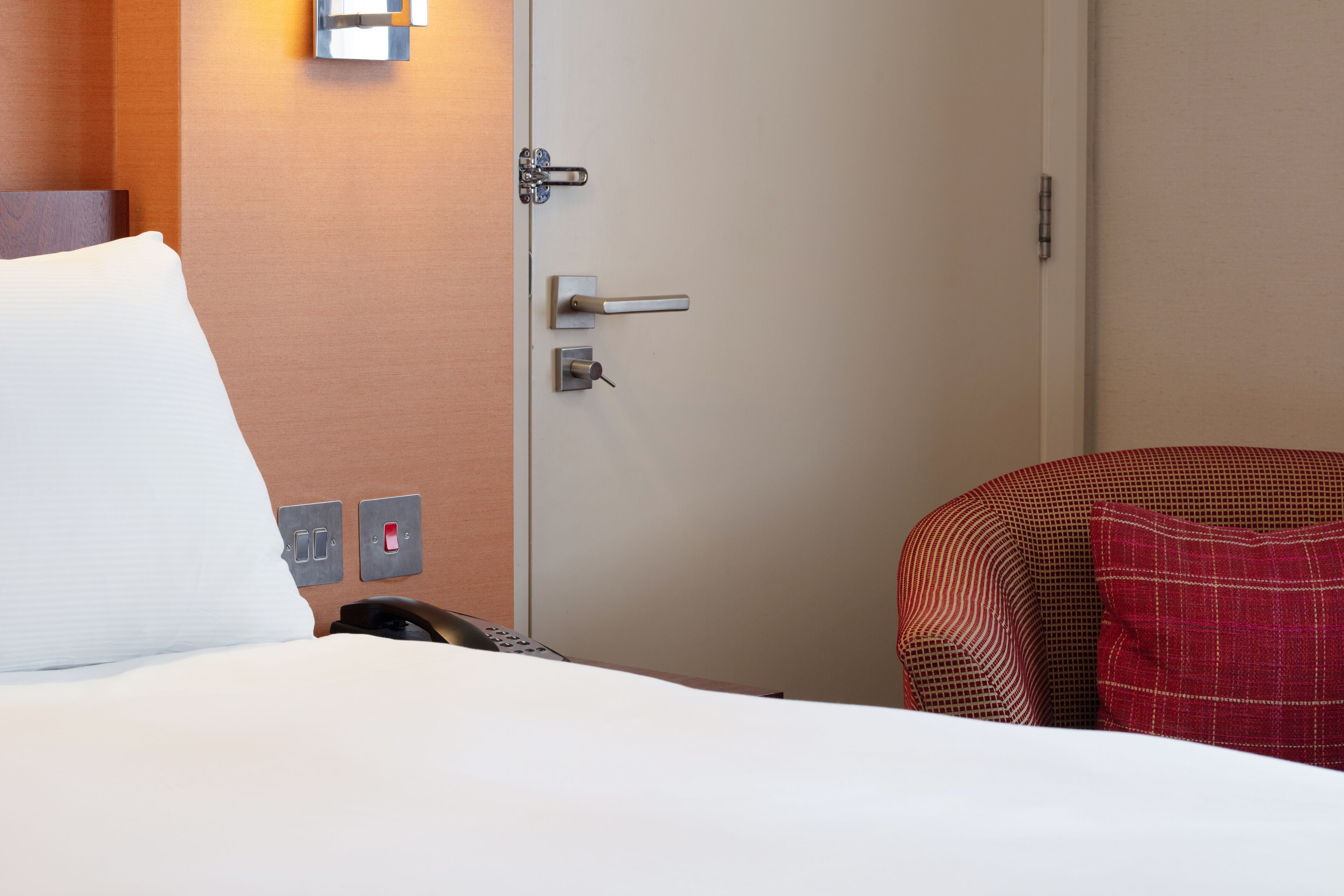 A hotel room with a bed, a door with a handle, a light switch, and a power outlet.