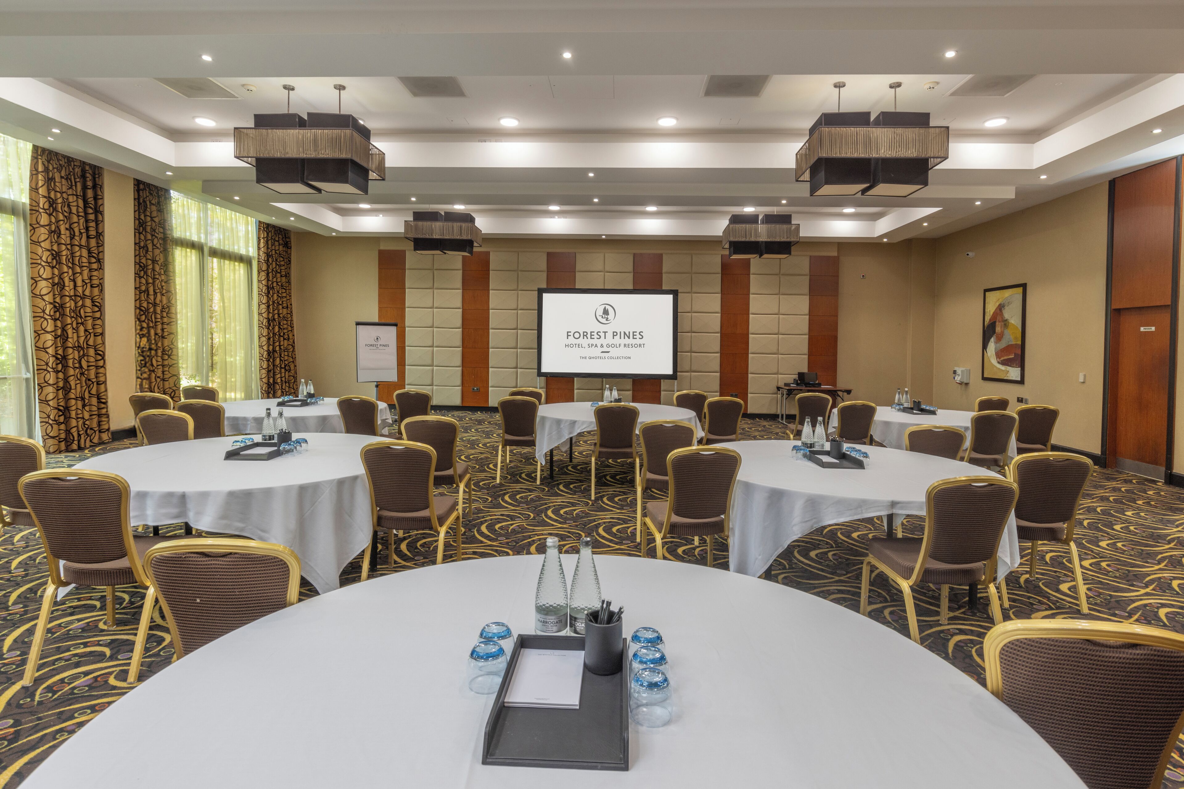 A conference room with multiple round tables, each with chairs and water bottles. A large screen displays the hotel's name.
