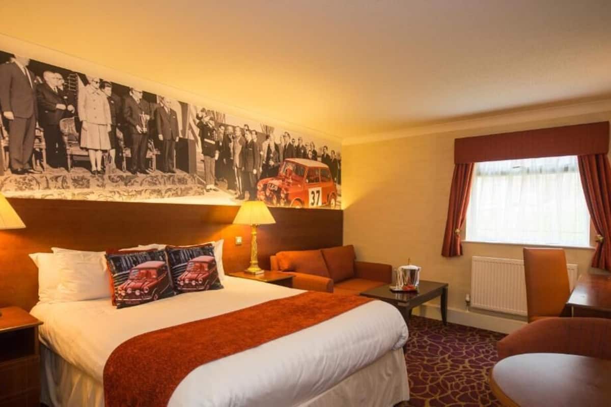 A hotel room with a large bed, two bedside lamps, a red throw blanket, a headboard with a black and white photo, a small table with a lamp, a sofa, a window with curtains, and a chair.