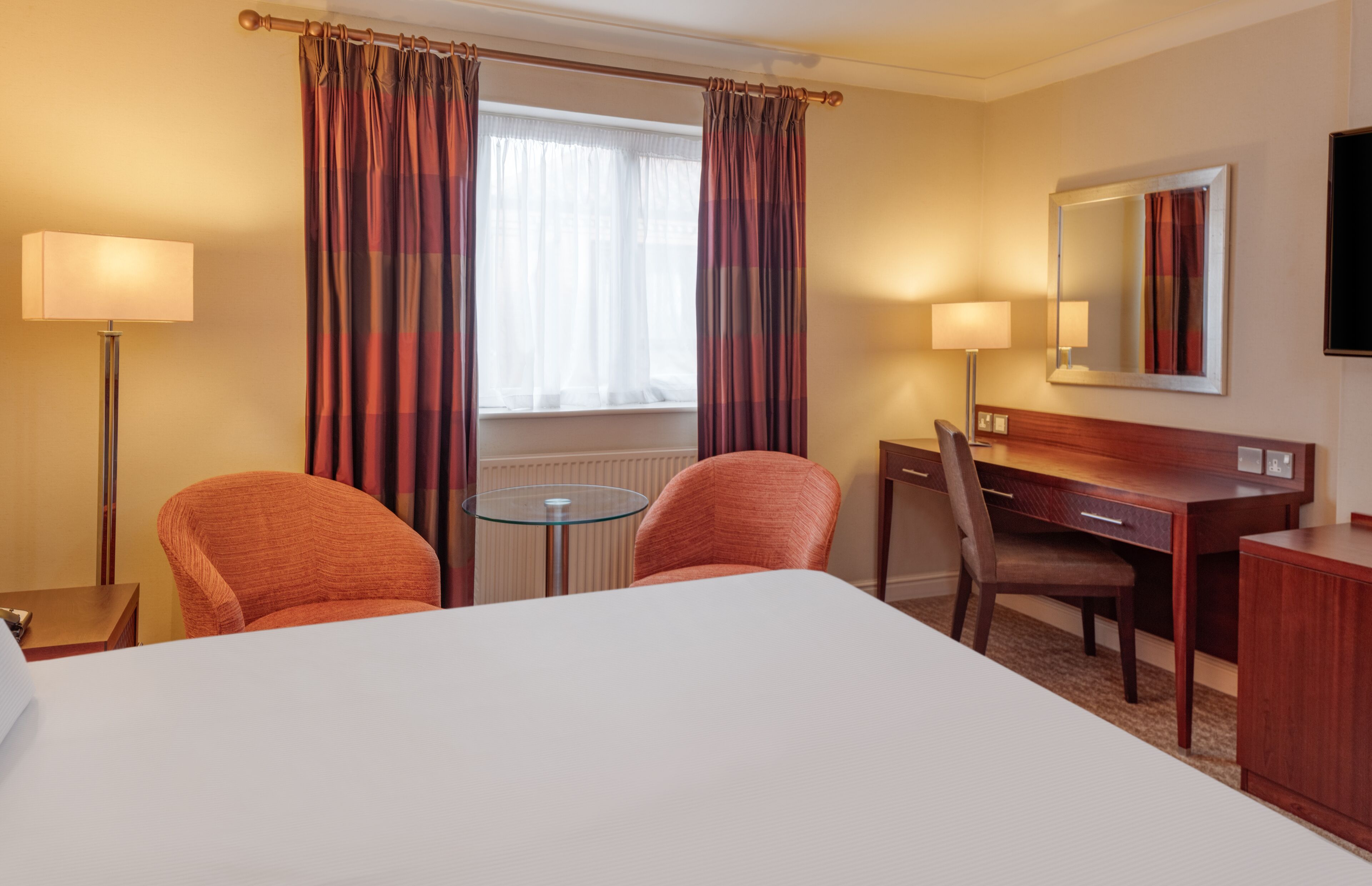 A hotel room with a large bed, two armchairs, a desk, a small round table, a lamp, a mirror, and a window with curtains.