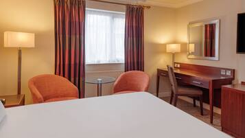A hotel room with a large bed, two armchairs, a desk, a small round table, a lamp, a mirror, and a window with curtains.
