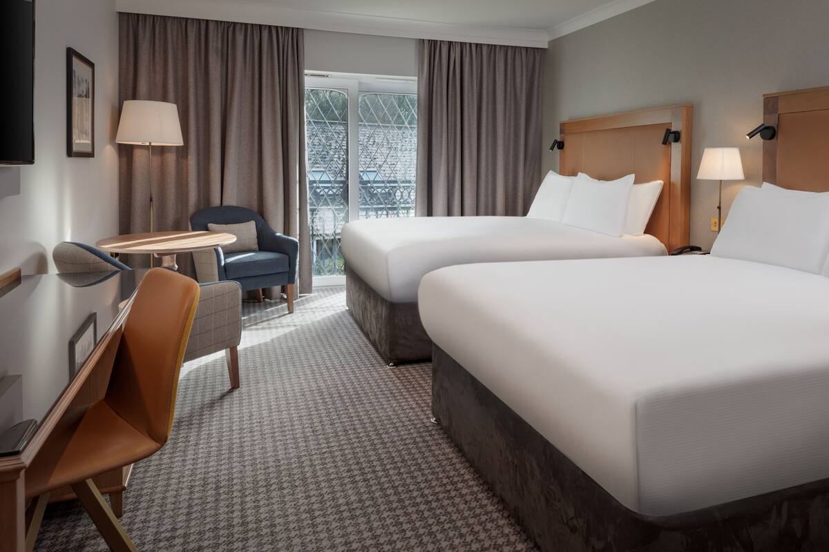 A hotel room with two beds, a desk, a chair, and a view of a building through large windows.