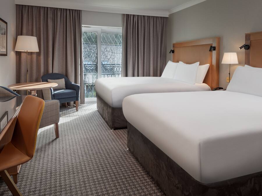 Une chambre d’hôtel avec deux lits, un bureau, une chaise et une vue sur un immeuble, visible à travers de grandes fenêtres.