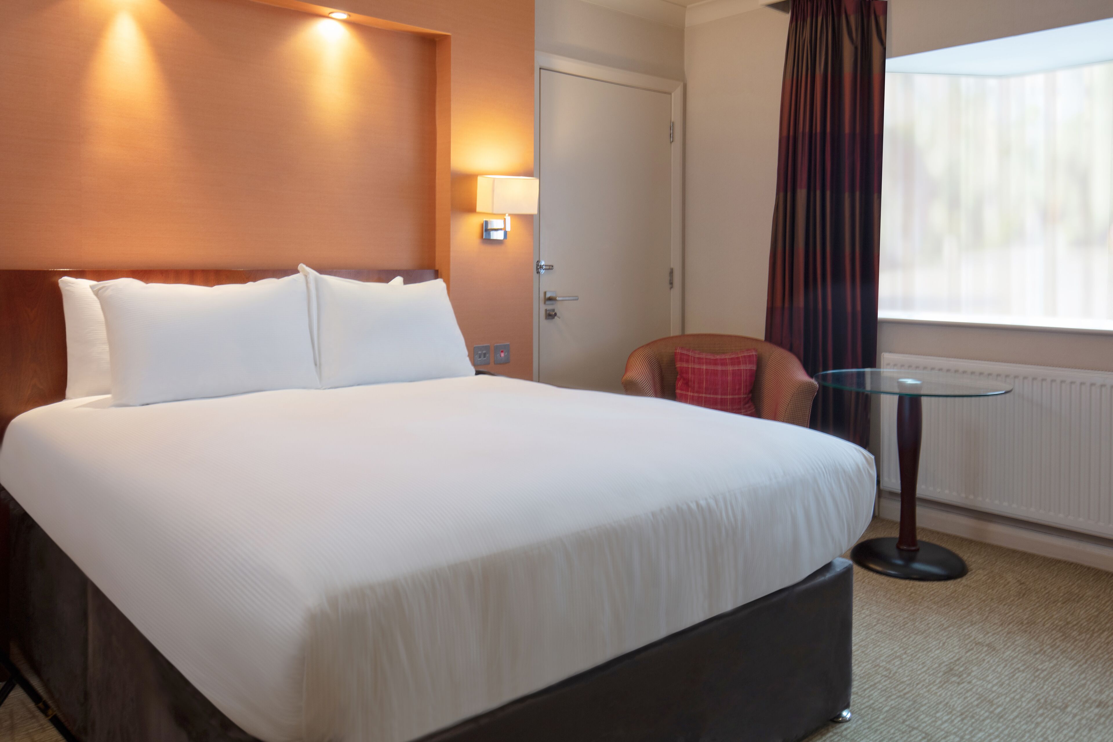 A hotel room with a large bed, a small round table, a chair, and a window with curtains.