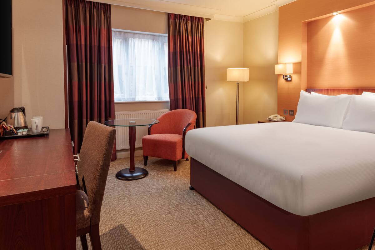A hotel room with a large bed, a desk, a chair, a table, and a lamp.