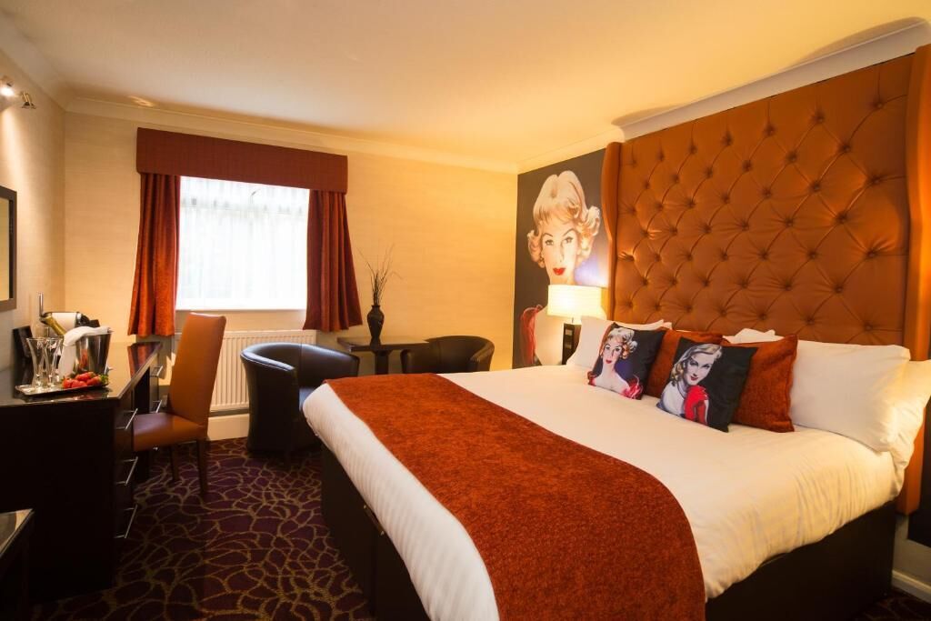 A hotel room with a large bed, a desk with a chair, a window with curtains, and a wall-mounted lamp.